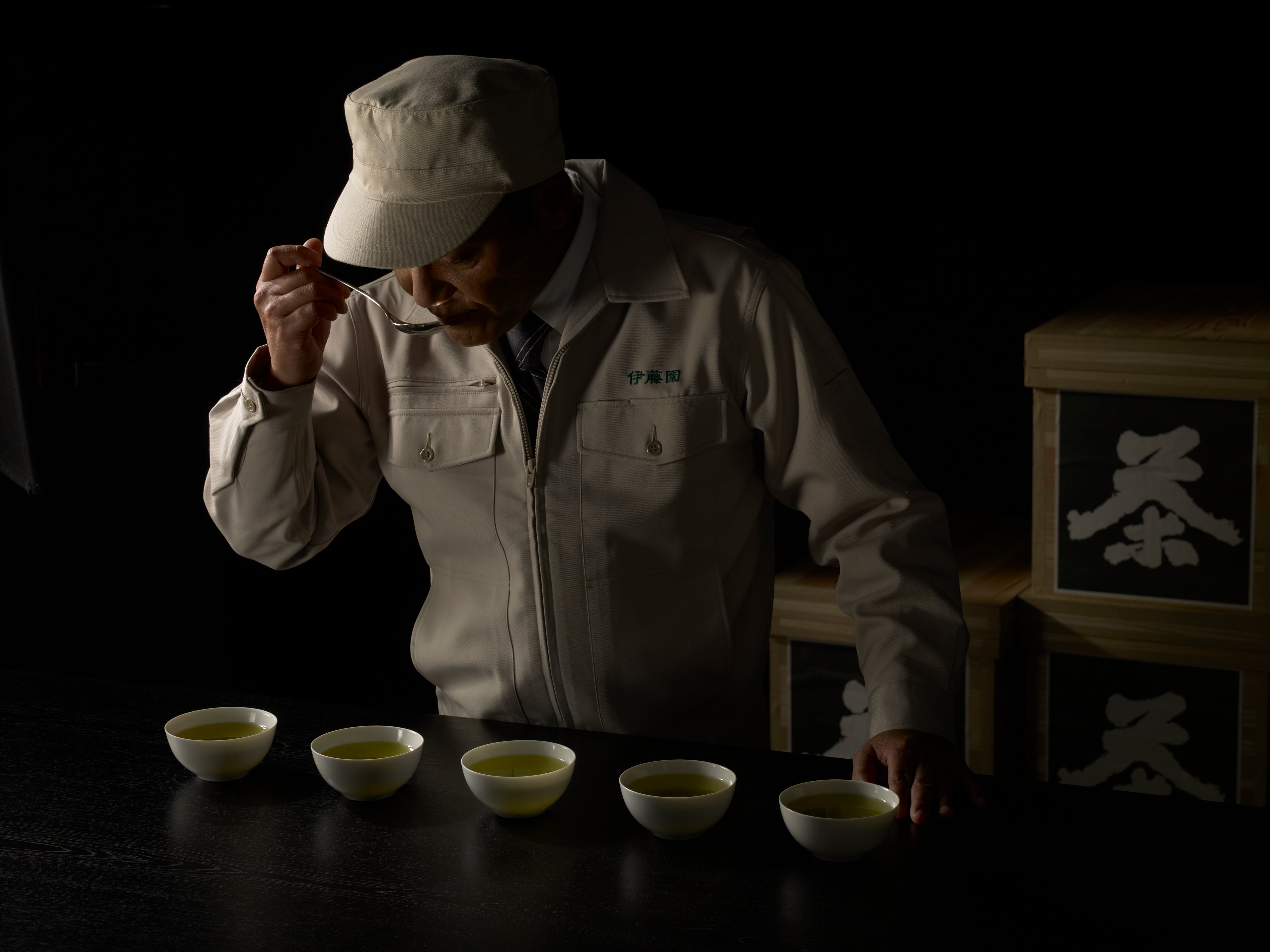 Person in a light jacket and cap tasting green tea from a row of five white bowls.