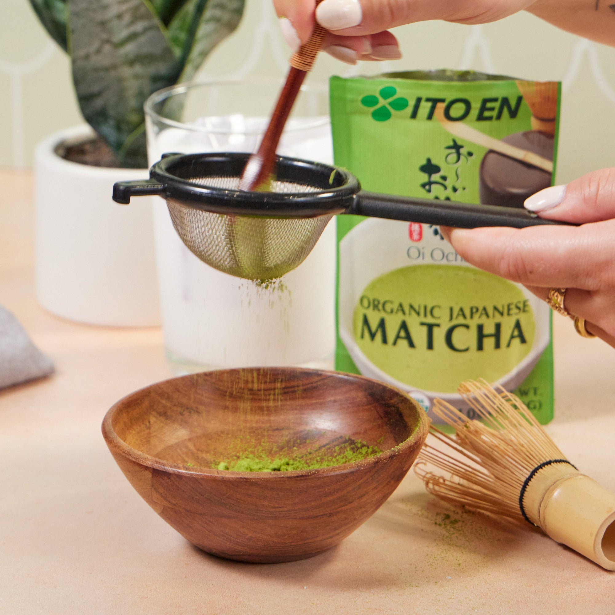 Hand holding sifter over a bowl of matcha, with a green plant and matcha pack in background.