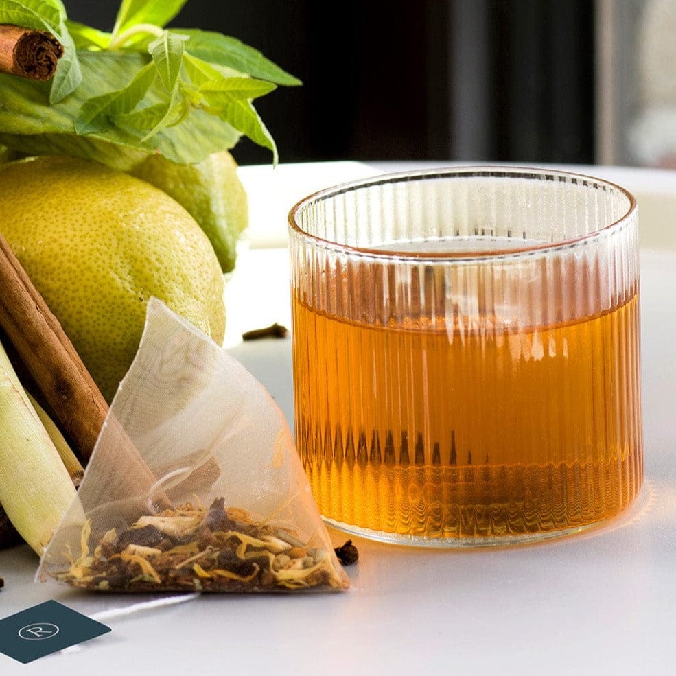 Clear mug of tea on a table with lemon, cinnamon, mint, and a tea bag.