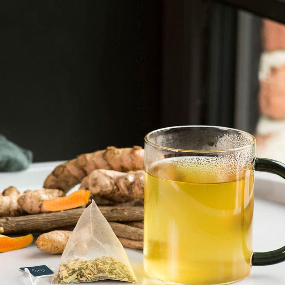 Glass mug with yellow tea beside ginger, orange slices, and tea bag on a white surface.