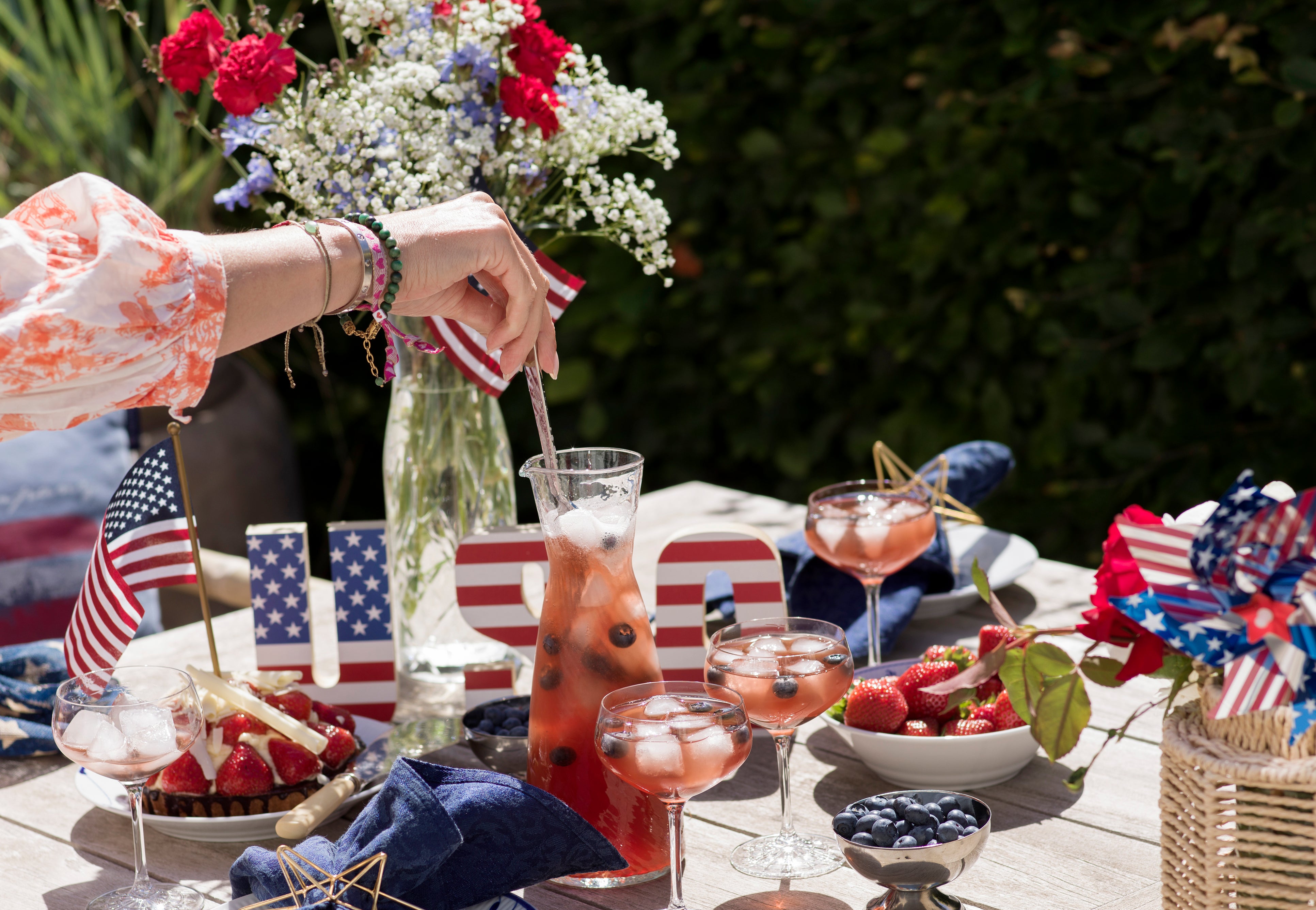 Person stirring iced drink with berries on a festive table with flowers and patriotic decor.