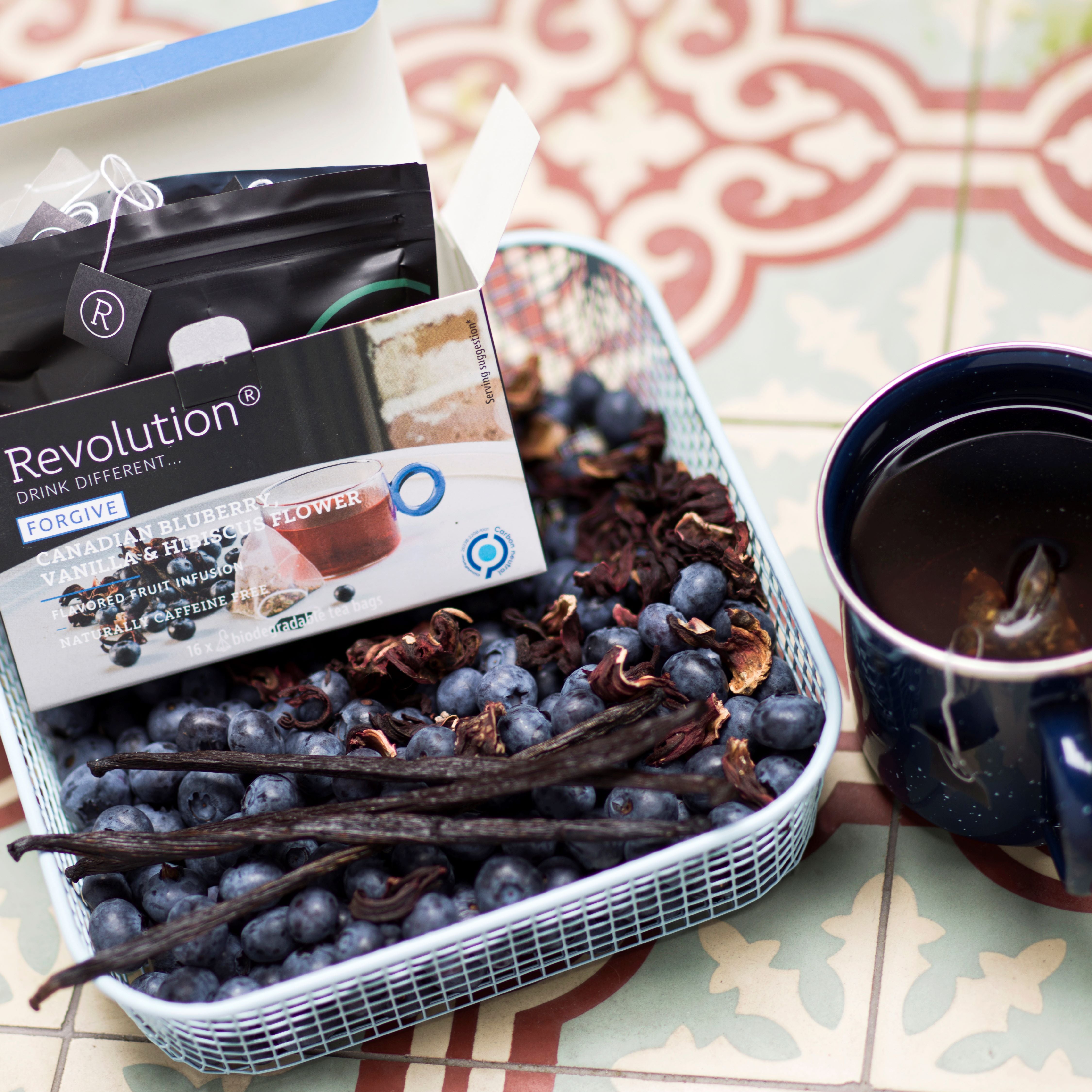 Tea box, cup, blueberries, and vanilla beans on a decorative surface.