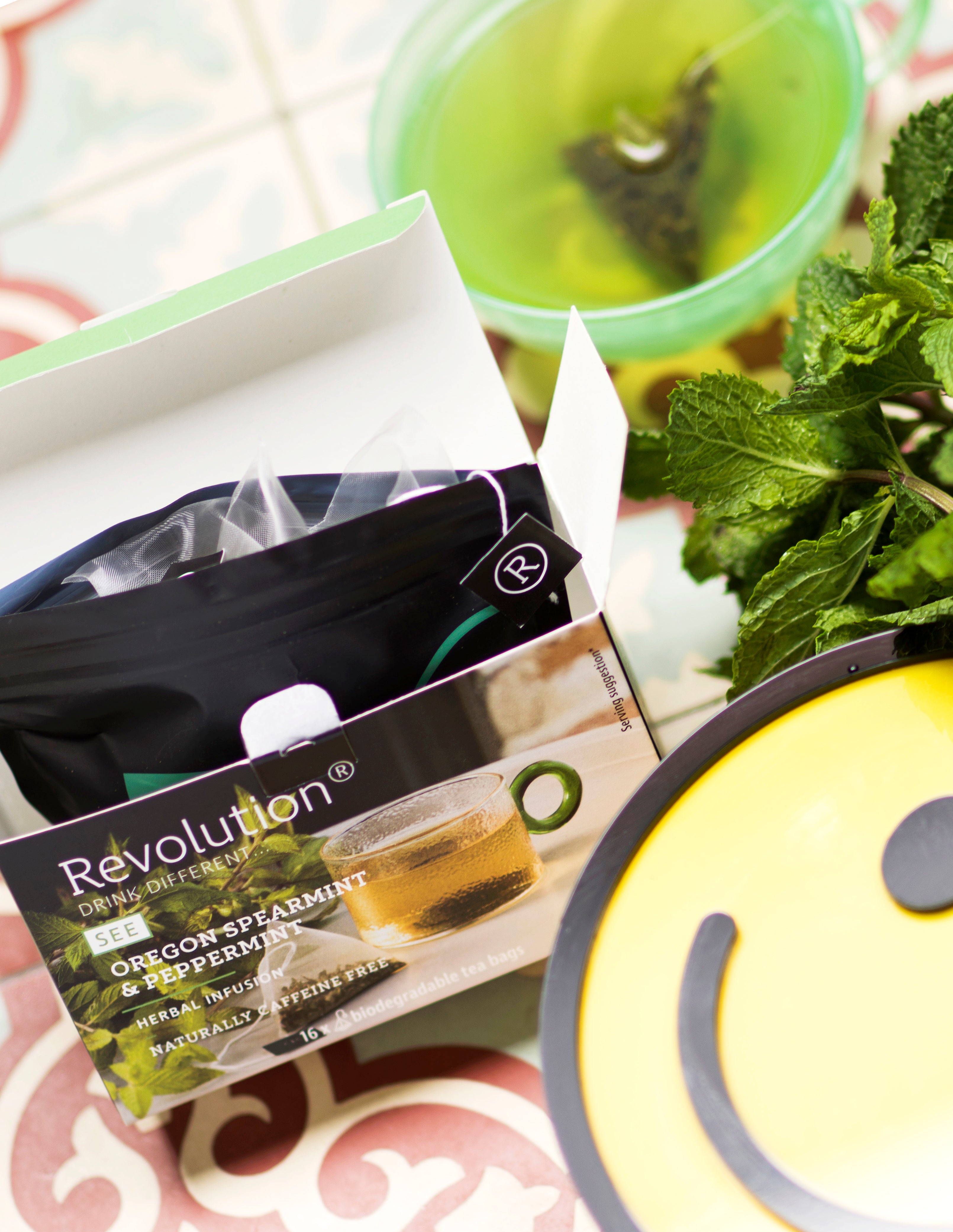 Tea box, green tea in a cup, mint leaves, and a smiley face plate.