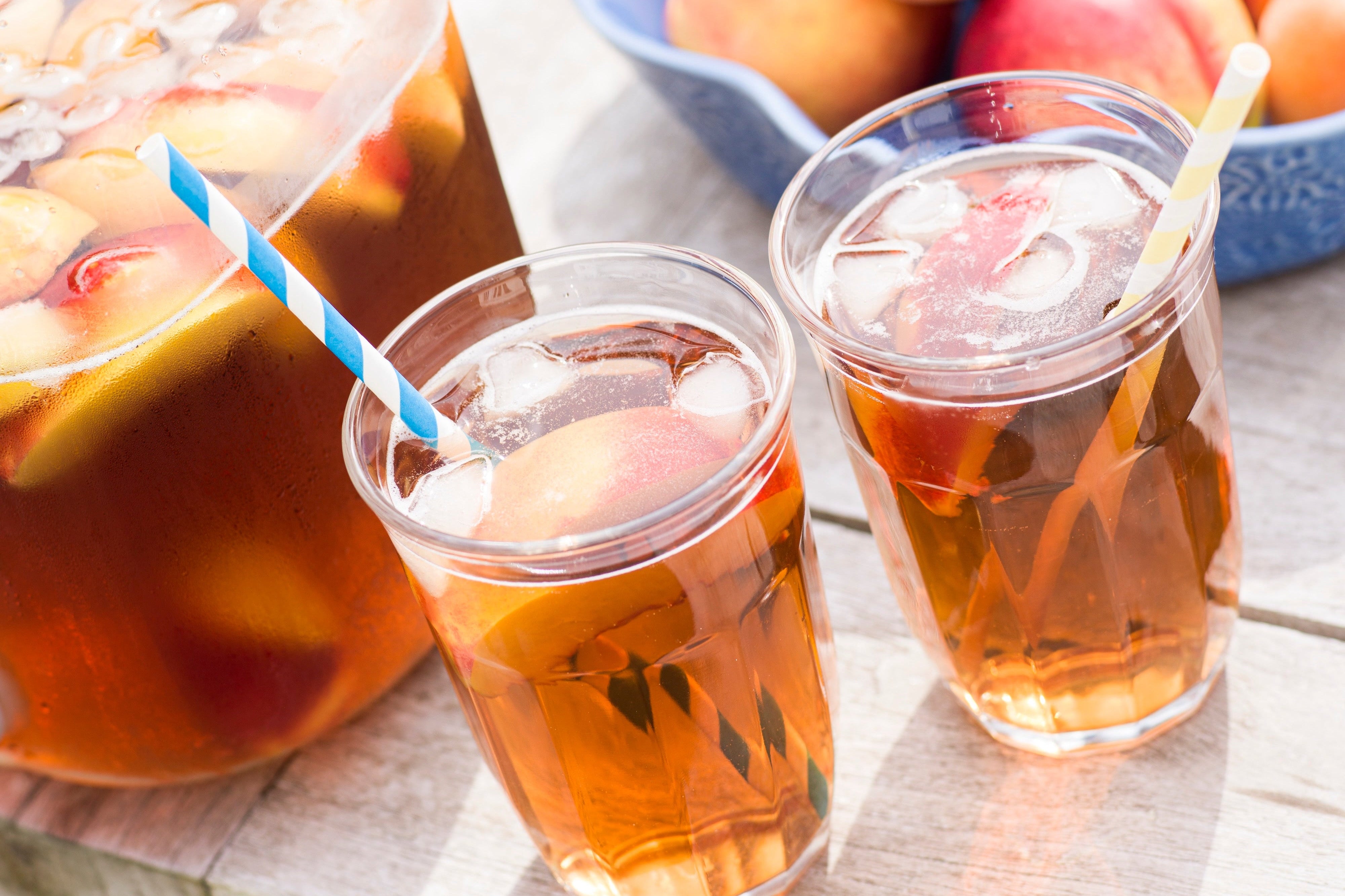 Peach iced tea in glasses with a large jar of peach iced tea and a bowl of peaches on a table.