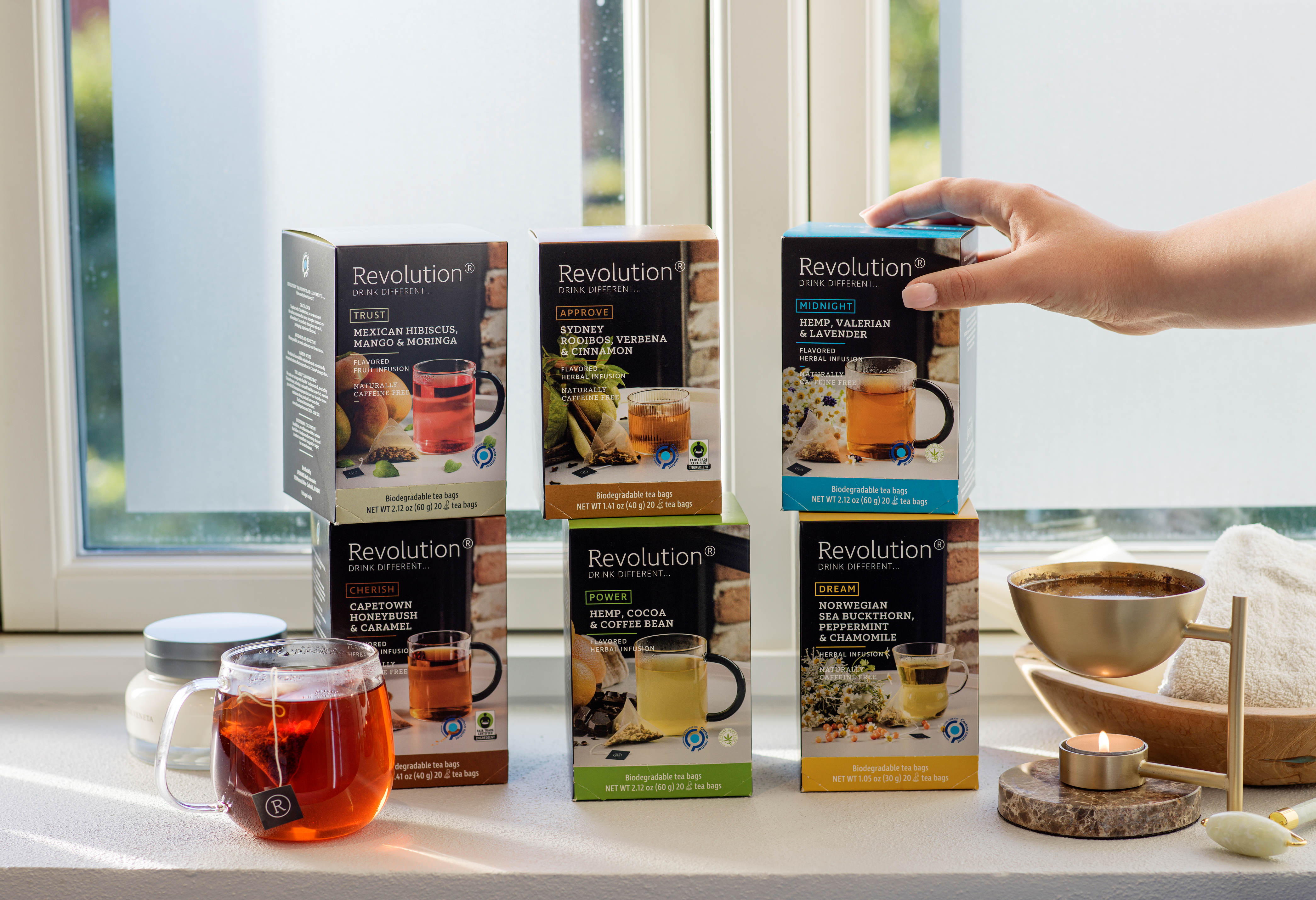Stack of tea boxes on a windowsill with a teapot, candle, and kitchen items.