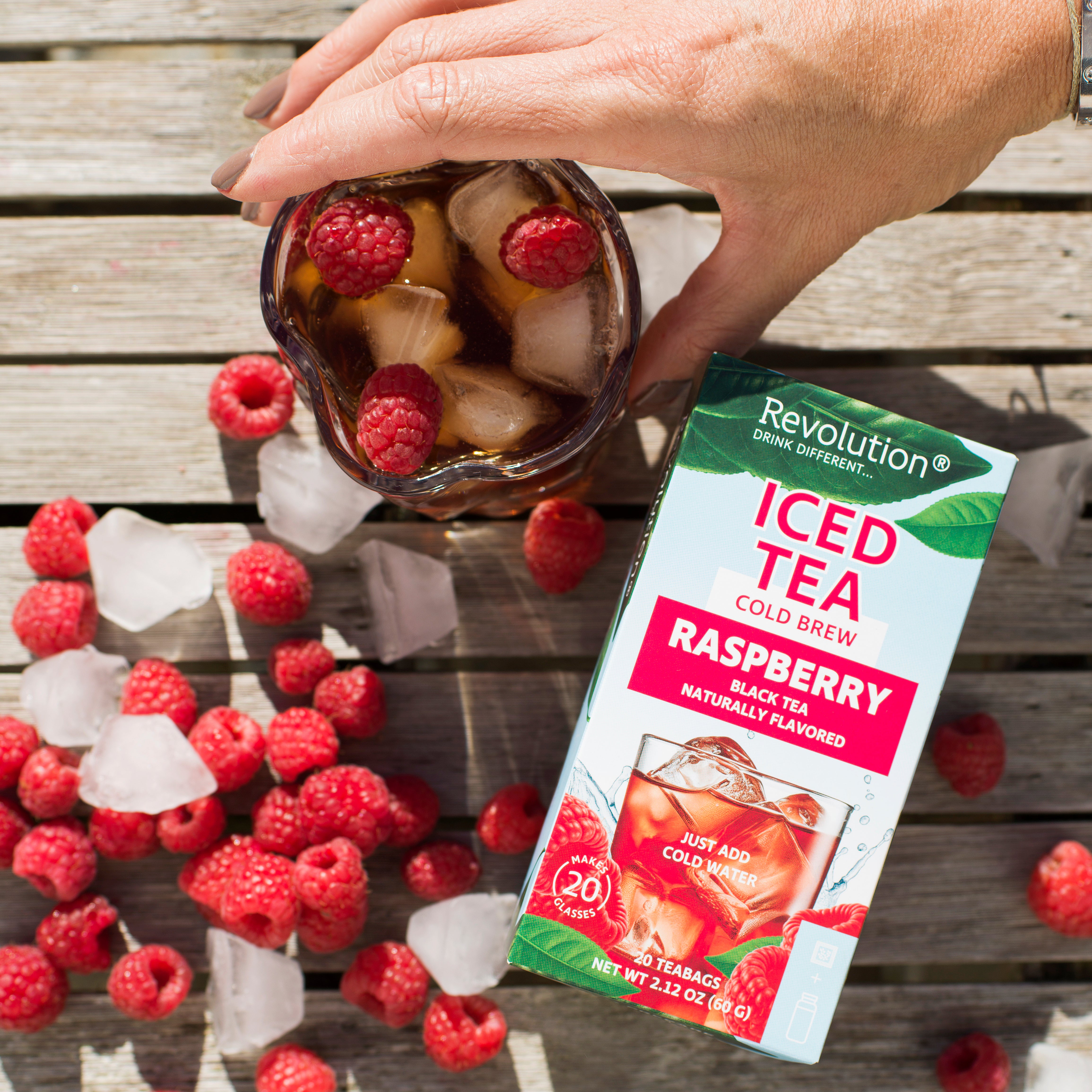 Hand holds iced tea with raspberries; iced tea box and scattered raspberries on a wooden surface.