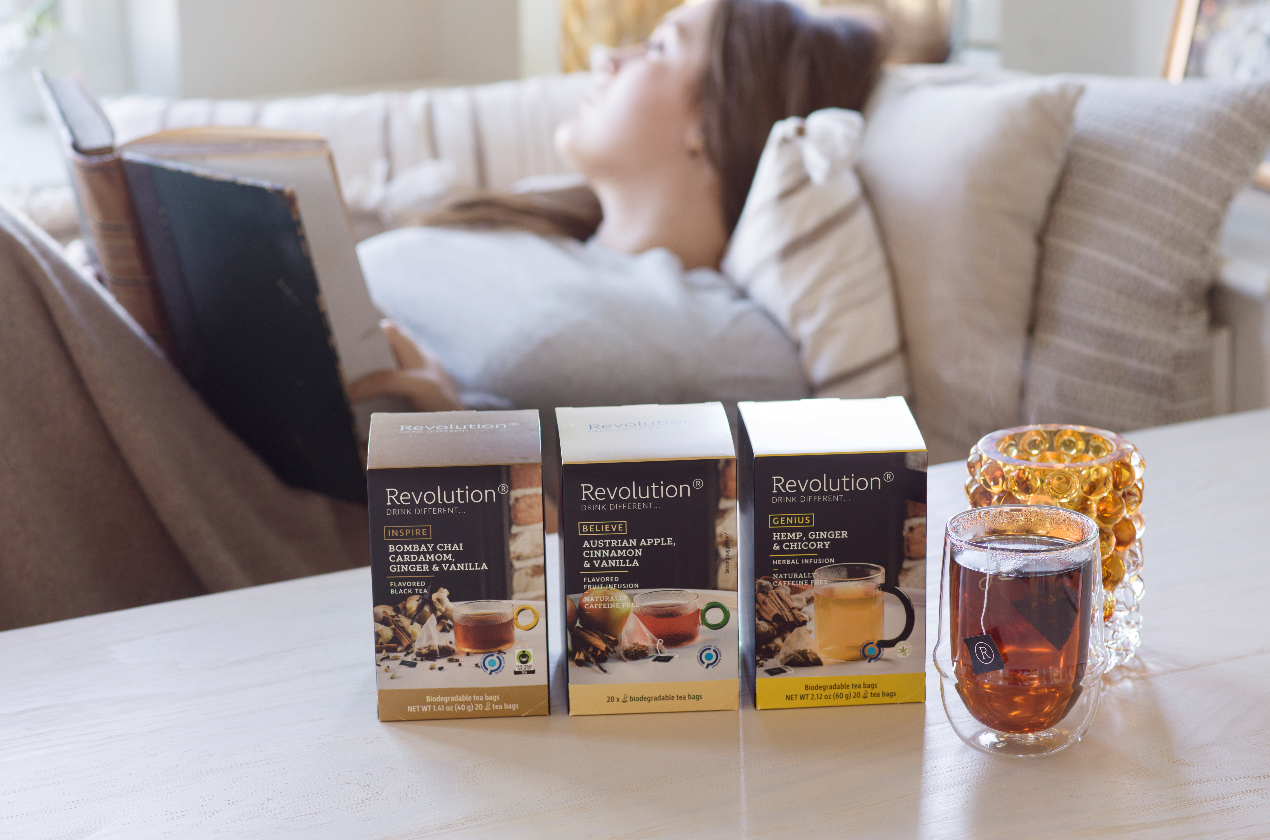 Person relaxing with tea and a book, next to three tea boxes and a glass of tea.
