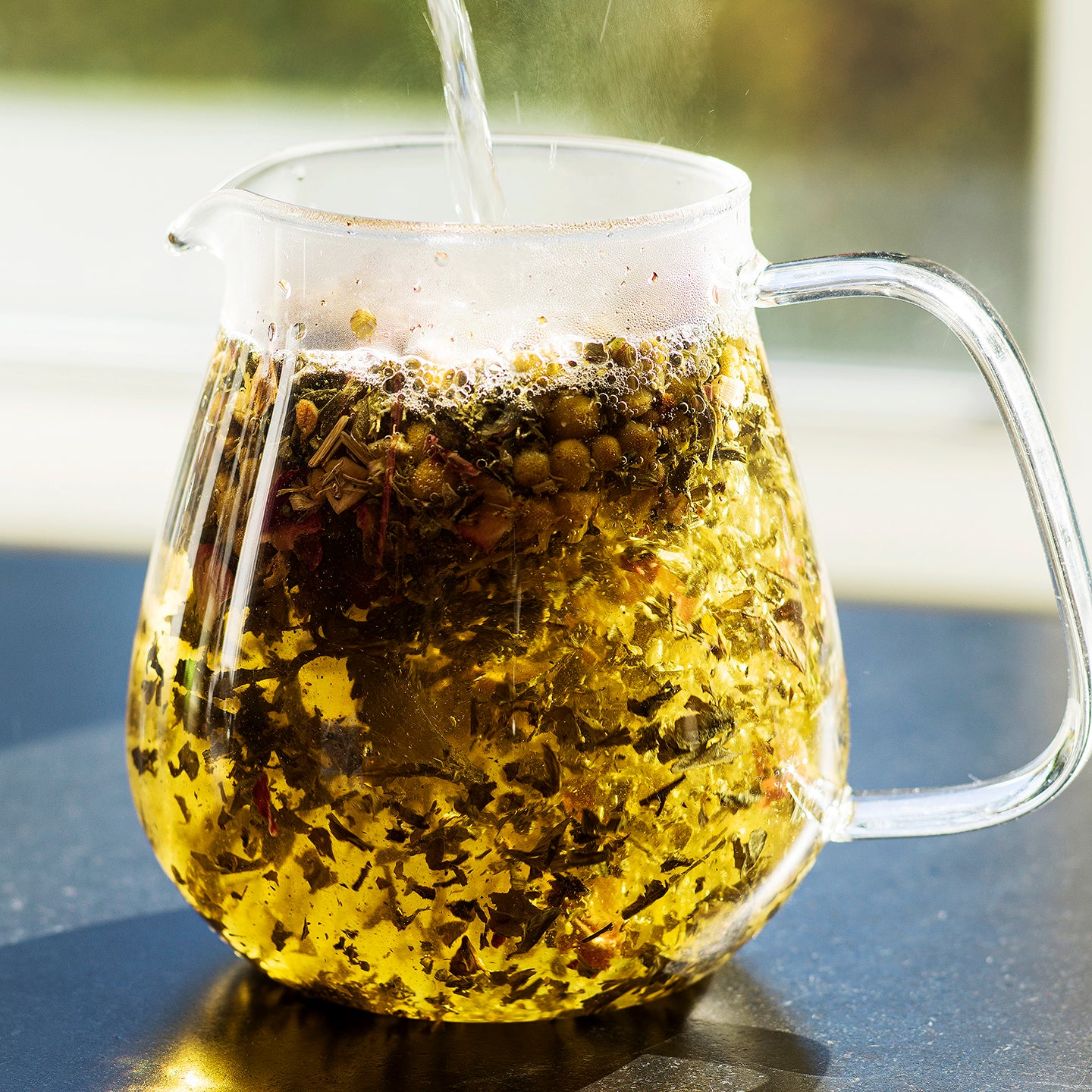 Clear pitcher of herbal tea steeping with visible herbs and flowers inside.