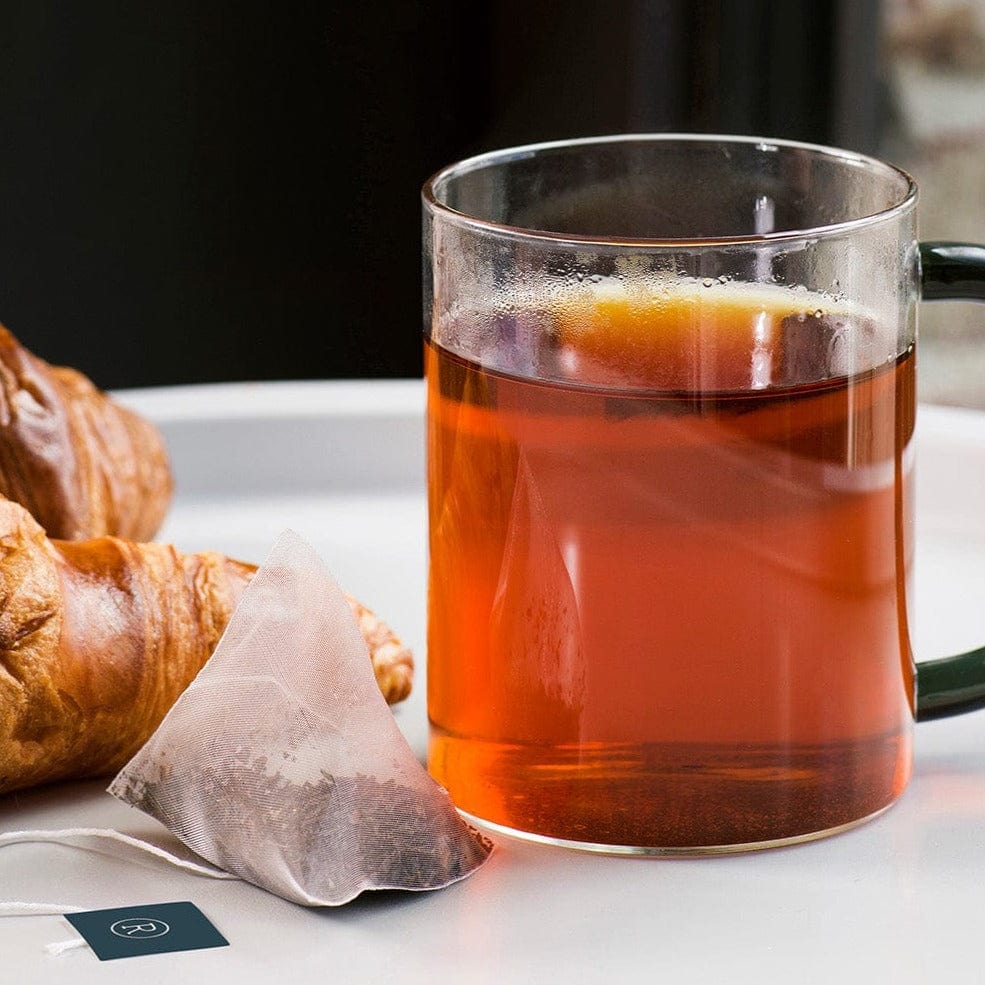 Glass mug of tea with a tea bag and croissant on the side on a white surface.