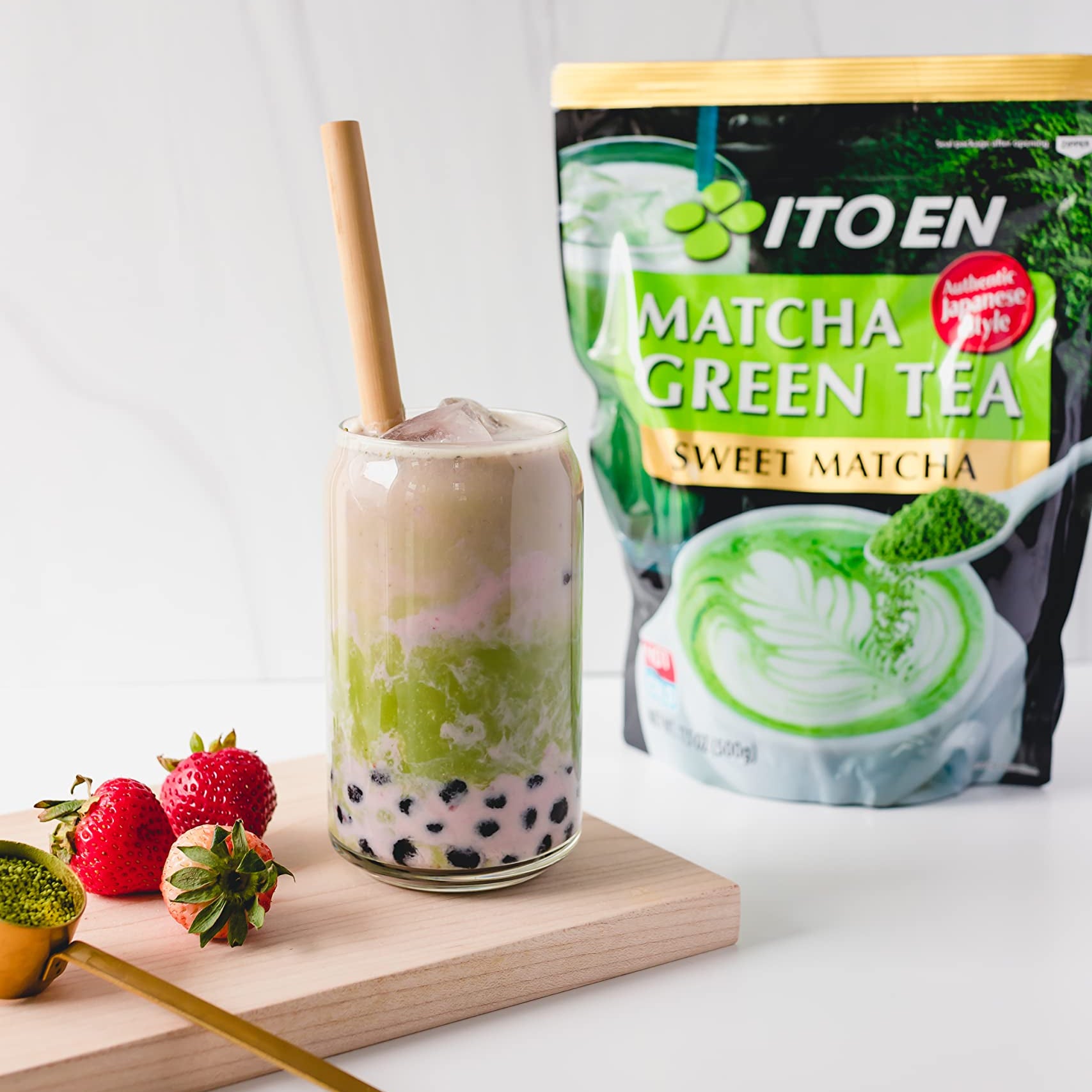 Layered matcha and milk boba drink with a straw, strawberries, and matcha pack on a board.