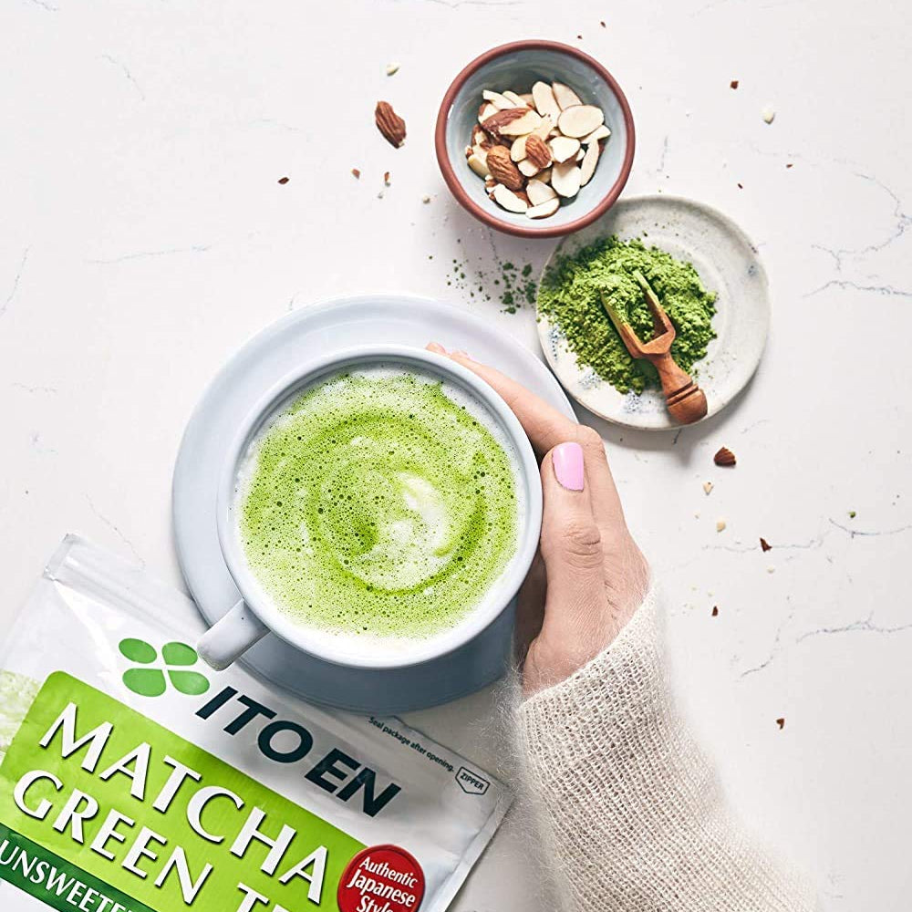 Hand holding matcha latte cup, with green tea pack, almonds, and matcha on white table.