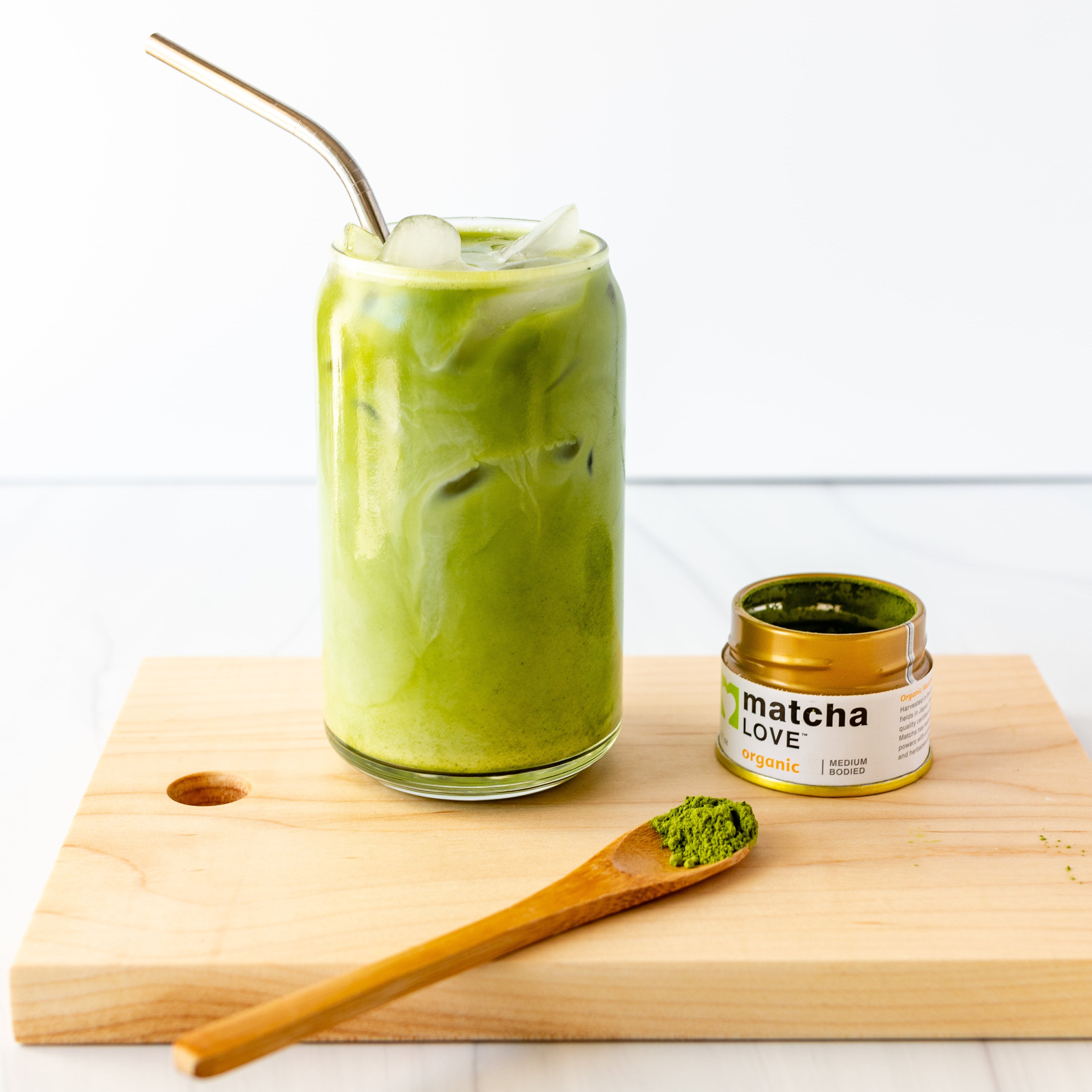 Iced matcha latte in a glass with a metal straw, next to an open matcha tin and a spoon on a wooden board.