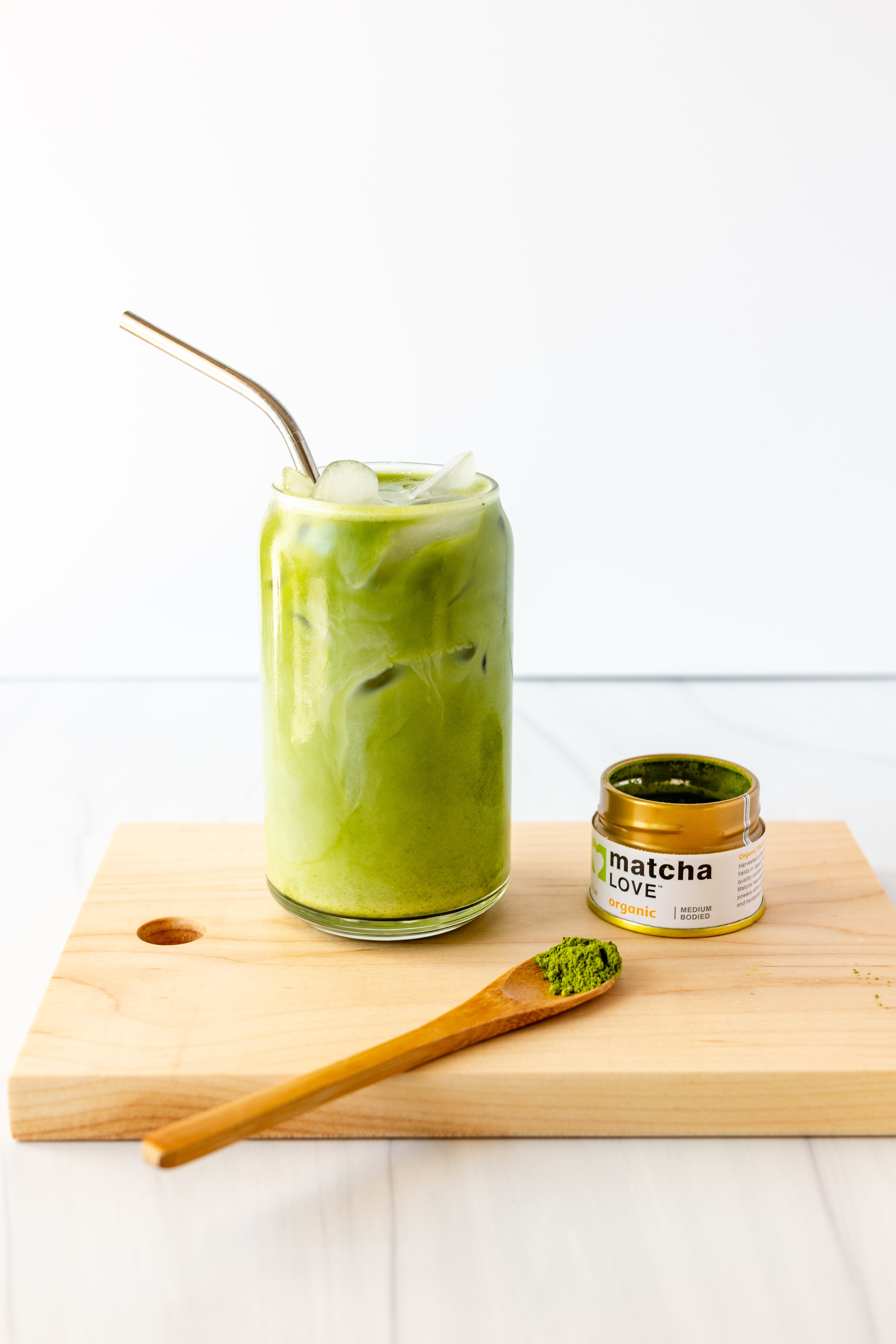Iced matcha drink with a metal straw, open matcha tin, and bamboo spoon on a wood board.