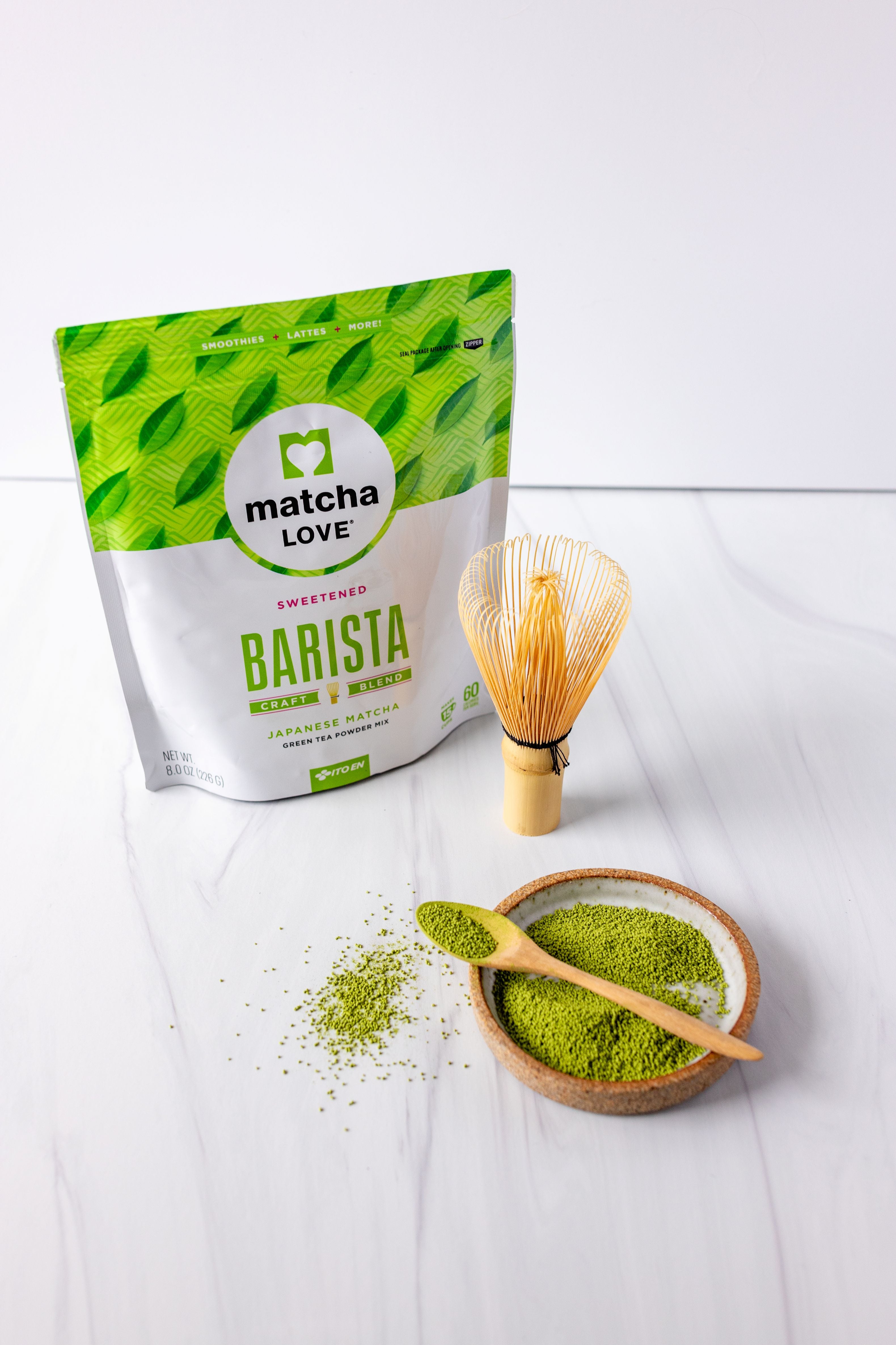 Matcha powder, bamboo whisk, and spoon sit beside a matcha bag on a white background.