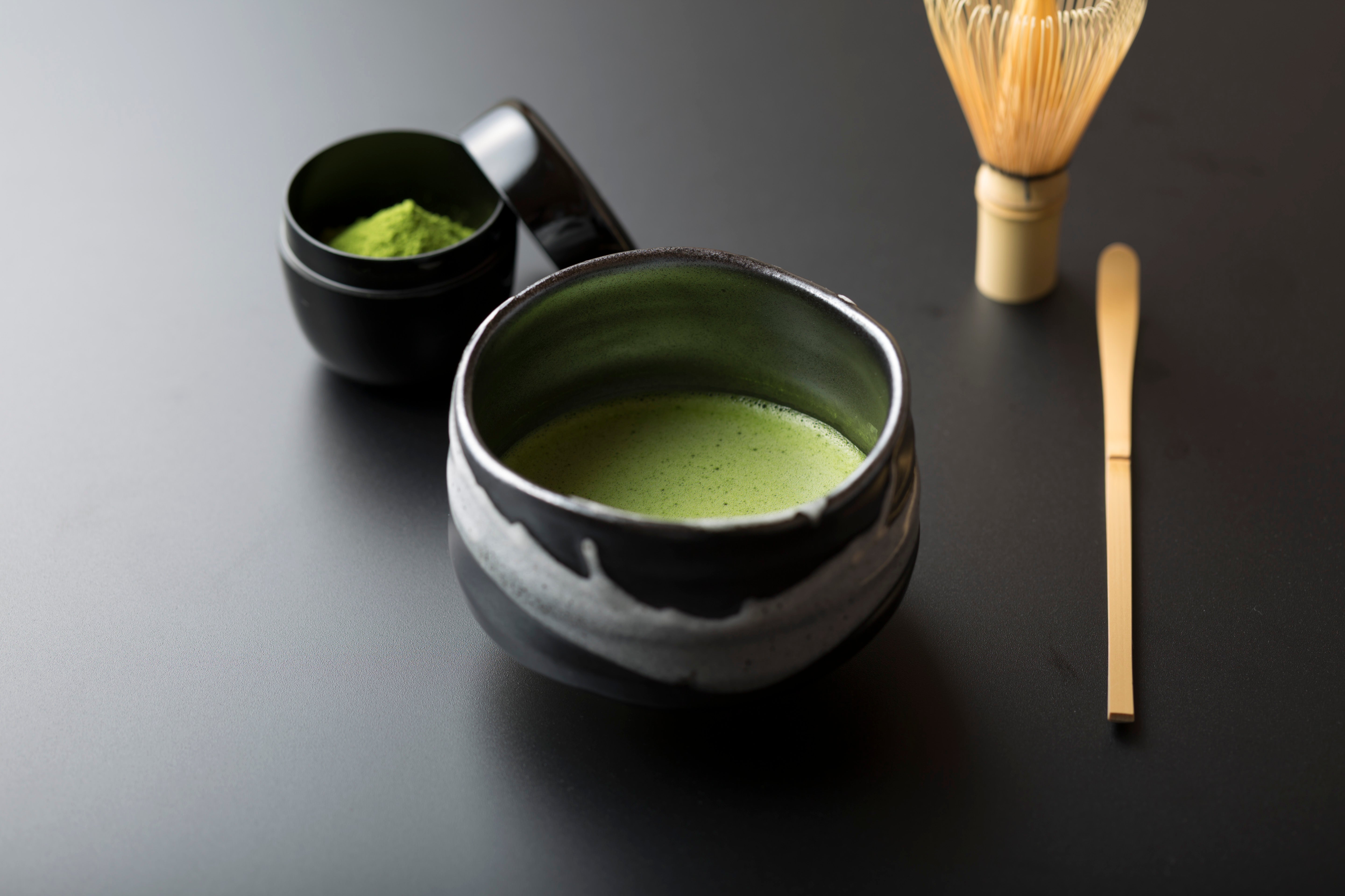 Ceramic cup of green tea on a black surface, with a whisk and a small black tea container.
