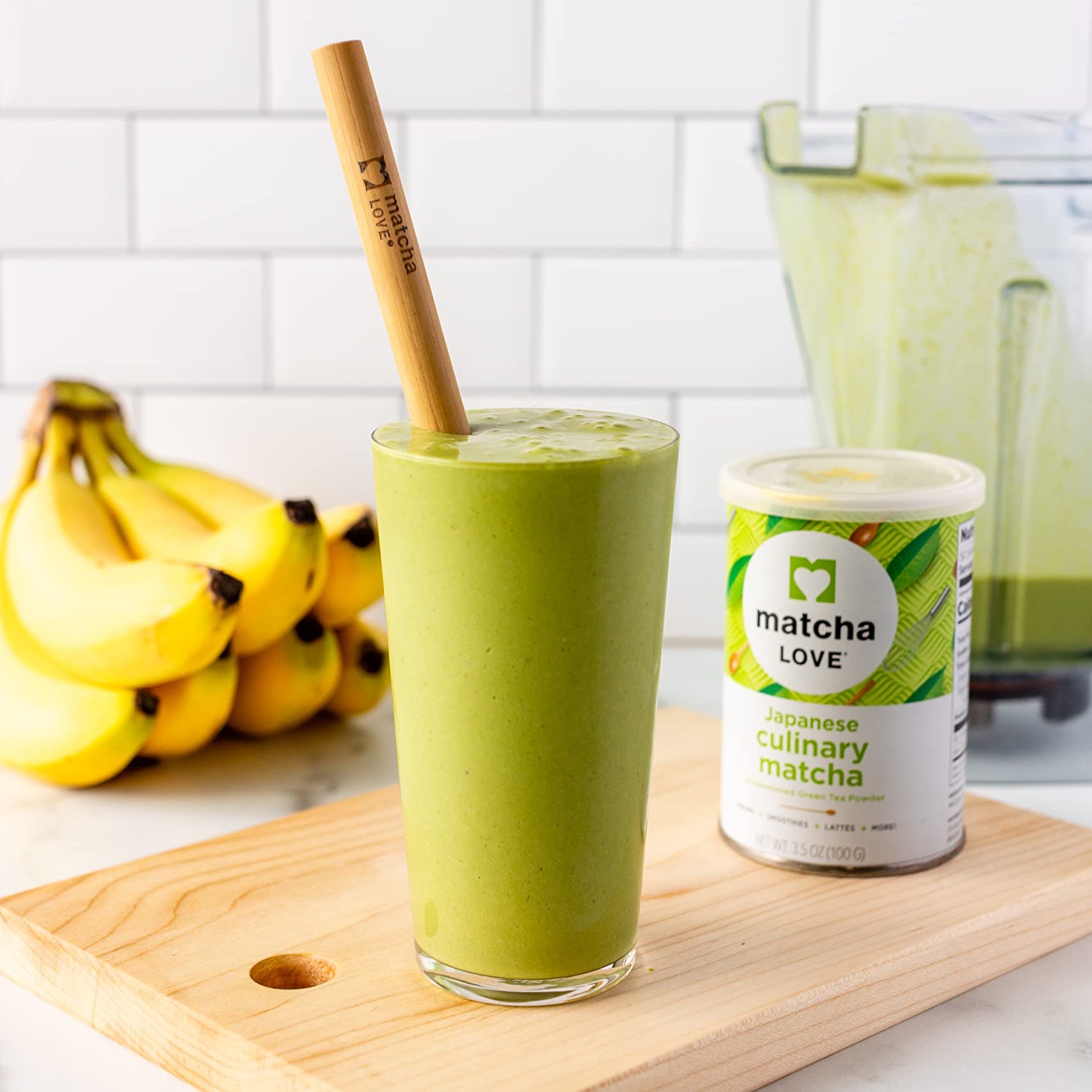 Green smoothie in glass with bamboo straw, next to matcha canister and bananas.