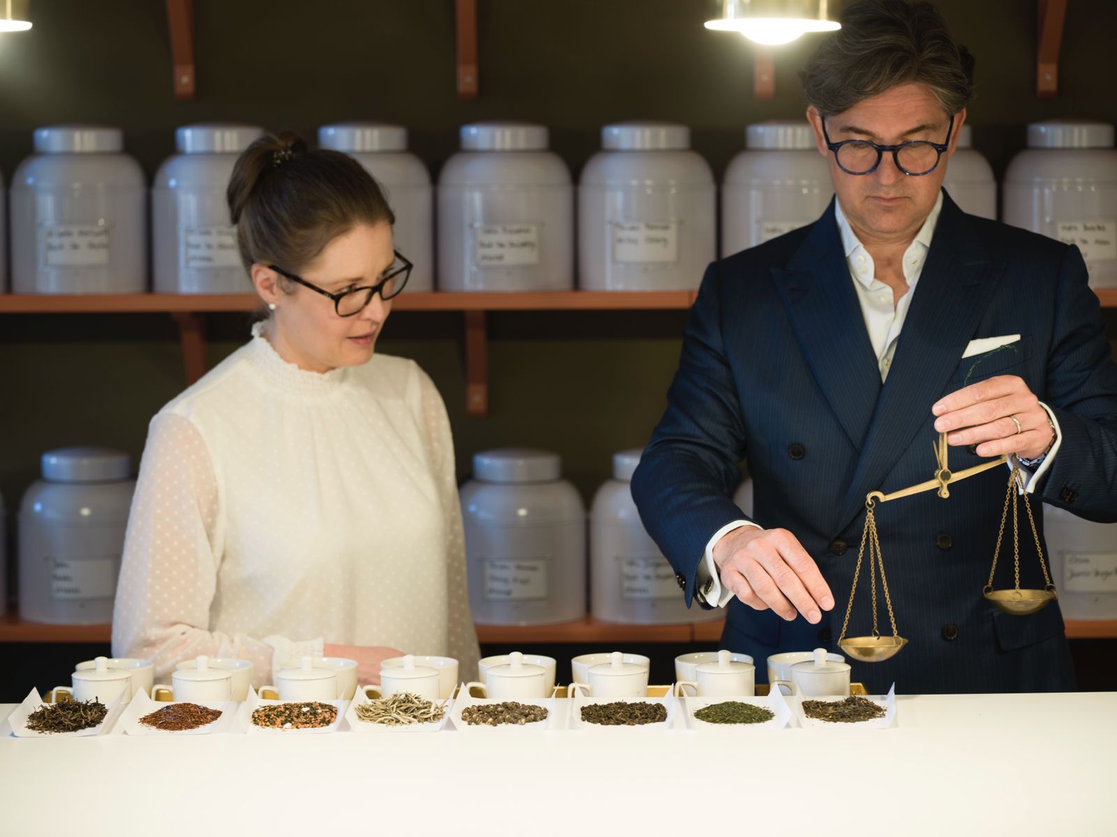 Man in blue suit weighing tea leaves on a scale while a woman in a white blouse watches.