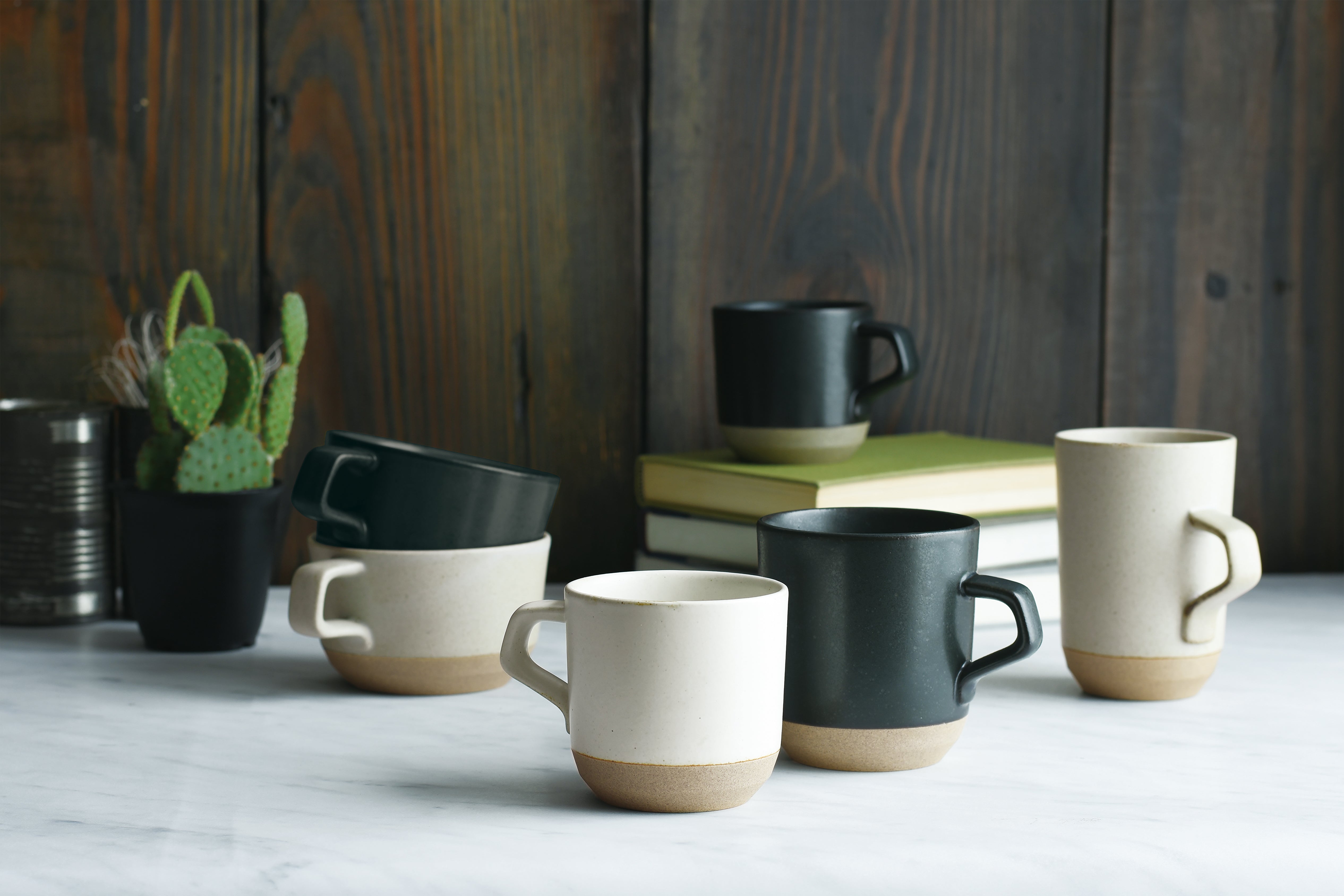 Six ceramic mugs in black and off-white on a marble surface with a potted cactus behind.