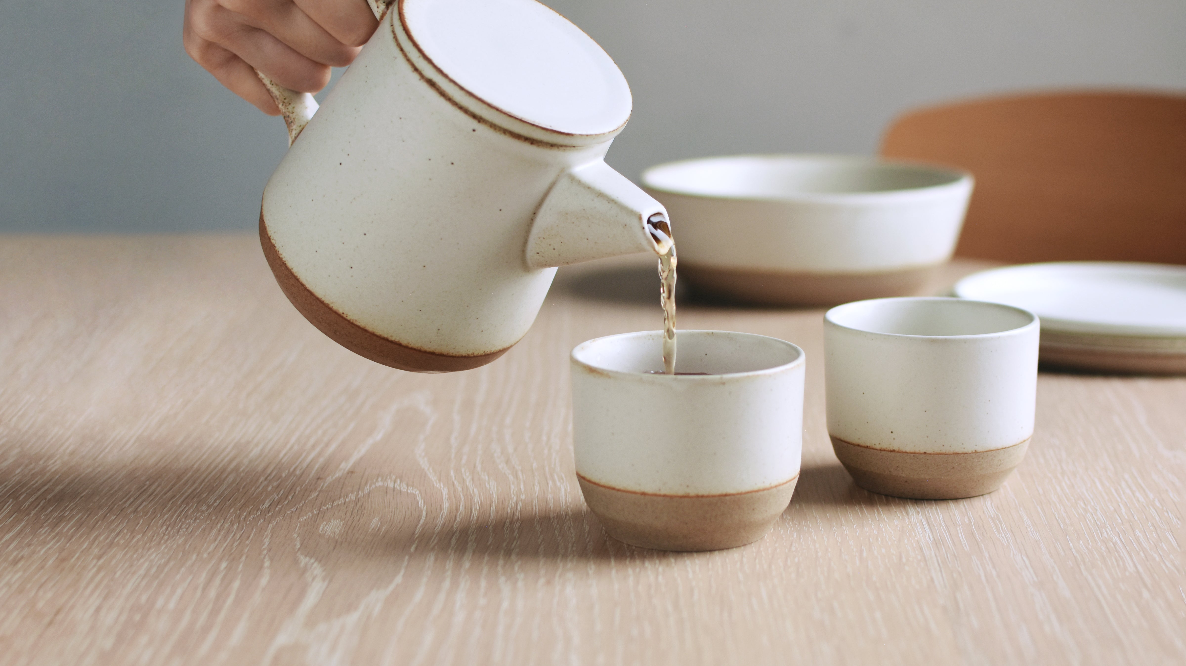 Hand pouring tea from a cream-colored teapot into a matching cup on a wooden table.